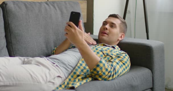 Joven hombre tiene un chat de vídeo en el teléfono inteligente, acostado en el sofá — Vídeo de stock
