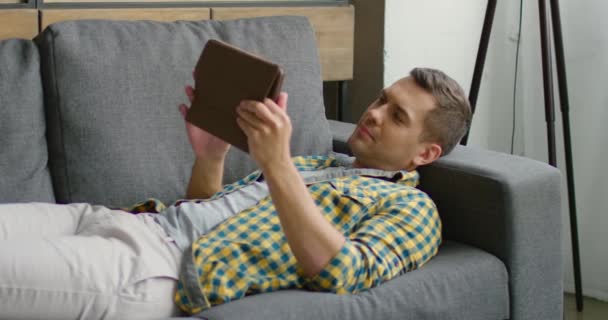 Ο νεαρός άνθρωπος είναι ξαπλωμένος στον καναπέ και περνά το χρόνο του στο διαδίκτυο, χρησιμοποιώντας τον υπολογιστή του tablet — Αρχείο Βίντεο
