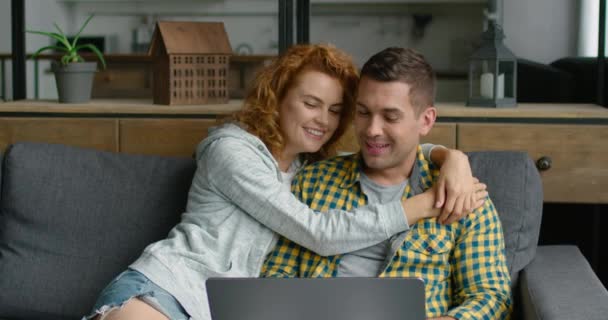 Young sweet couple is sitting on a sofa, browsing in laptop, talking about life — Stock Video