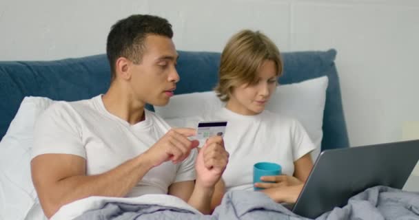若い混合レースのカップルは、ベッドに横たわって、オンラインショッピングフォームを埋めます — ストック動画