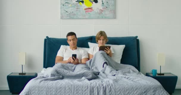 Смешанная раса пара лежит в постели, просматривая в гаджетах — стоковое видео