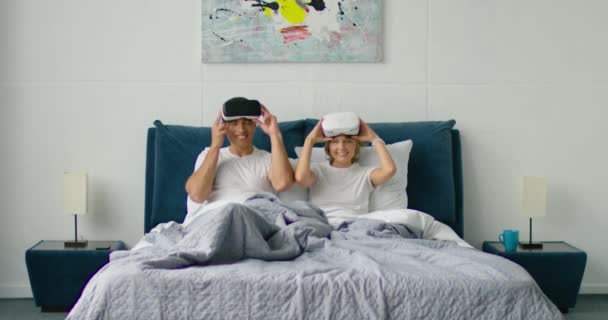 若い混合レースのカップルは、ワイヤレスヘッドセットを置いて、ベッドに横たわっています — ストック動画