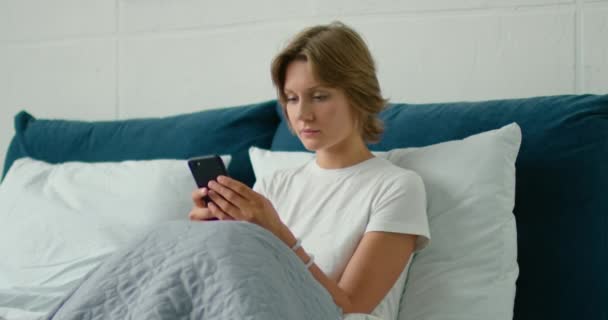 Jeune femme couchée au lit, textos dans son smartphone — Video