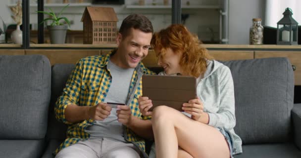 Młoda para lubi zakupy online, przy użyciu komputera typu Tablet, siedząc na kanapie — Wideo stockowe