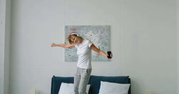 若い女性は、ワイヤレスヘッドセットを着用して、ベッドの上でジャンプして踊っています — ストック動画