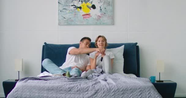 Молода змішана пара дивиться телевізор у спальні, людина в поганому настрої — стокове відео