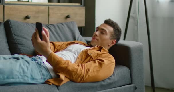 Hombre hispano guapo está acostado en el sofá, usando su teléfono inteligente — Vídeo de stock