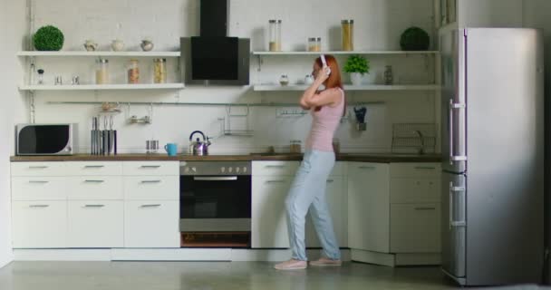 Hübsche Frau setzt drahtloses Headset auf, steht in der Küche — Stockvideo