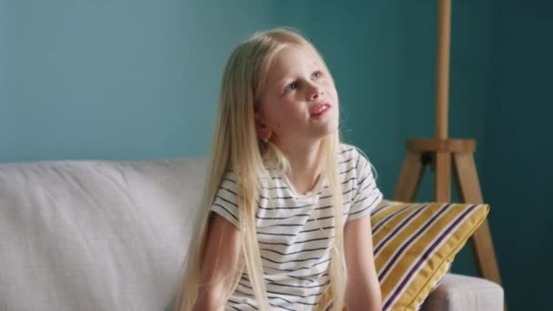 Menina loira pequena está sentada no sofá e pensando — Vídeo de Stock