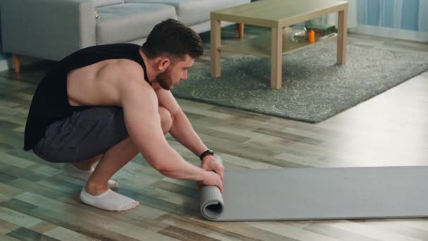 Hombre joven está rodando encima de la alfombra de fitness y mira a la cámara — Vídeos de Stock