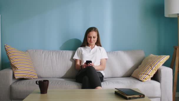 Красивая женщина использует мобильный телефон, сидя на диване дома — стоковое видео