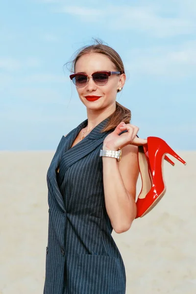 Portrait of stylish white young woman in suit, sunglasses, red lipstick standing with red glossy high heels shoes in her hands outdoor in desert