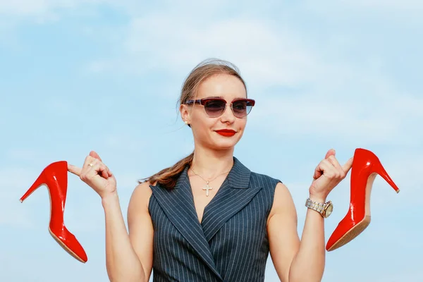 Portrait of stylish white young woman in suit, sunglasses, red lipstick standing with red glossy high heels shoes in her hands outdoor in desert