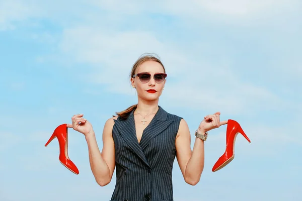 Portrait of stylish white young woman in suit, sunglasses, red lipstick standing with red glossy high heels shoes in her hands outdoor in desert