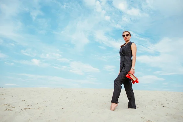 full height portrait of stylish white young woman in suit, sunglasses, red lipstick standing with red glossy high heels shoes in her hands outdoor in desert