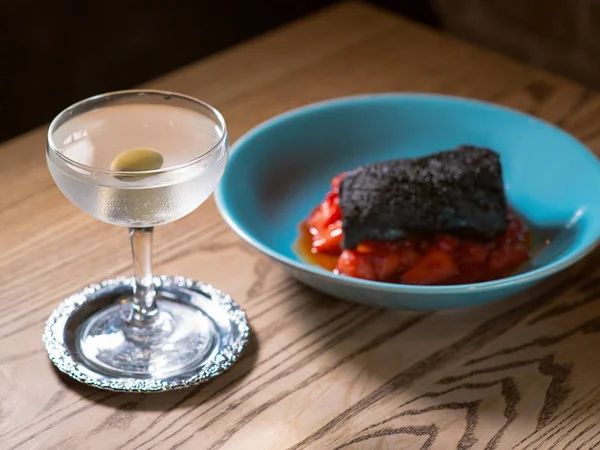 baked pork tenderloin marinated in cuttlefish ink with red tomatos and basil on blue plate and dry vermouth alcohol cocktail with olive on the table in the bar