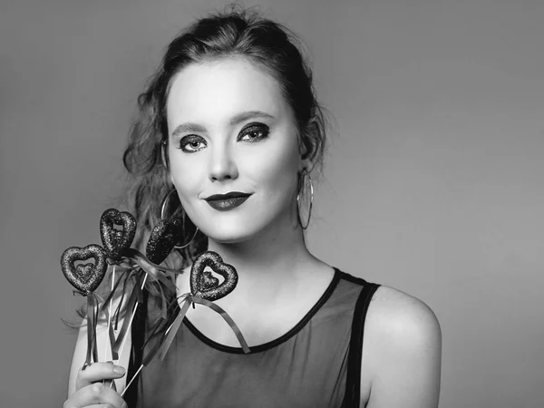 monochrome portrait of beautiful attractive stylish sad young redhead woman in earrings and with make up with saint valentine\'s day decorations (hearts) in her hands