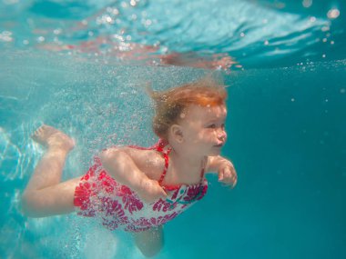 Mavi yüzme havuzunda sevimli modern elbise dalış sualtı gülümseyen bebek kız. Aktif yaşam tarzı, ebeveynlerle çocuk yüzme dersi. Tatil sırasında aile yaz tatili sırasında su sporları aktivitesi