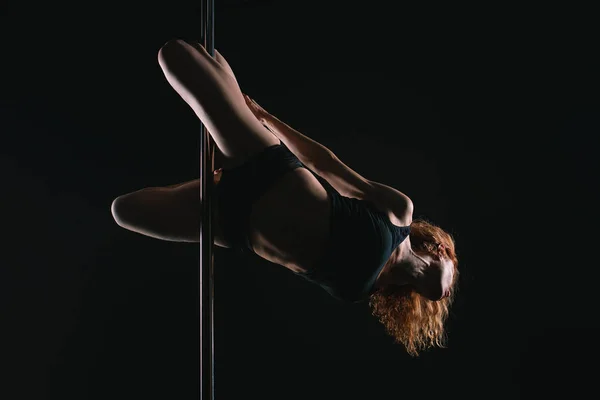 Pole sports: beautiful young athletic fit strong redhead woman gymnast in black T-shirt and shorts doing sport exercises (training, work out) on the pole on dark background