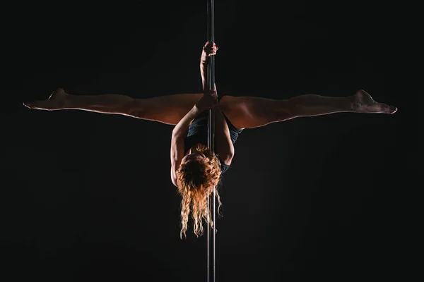 Pole sports: beautiful young athletic fit strong redhead woman gymnast in black T-shirt and shorts doing sport exercises (training, work out) on the pole on dark background