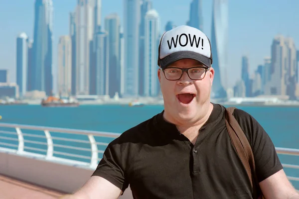 portrait of Impressed by modern Dubai (wow effect) european caucasian redhead man in glasses and hat travel to United Arabic Emirates on the cityscape, skylines and sea background in sunny day