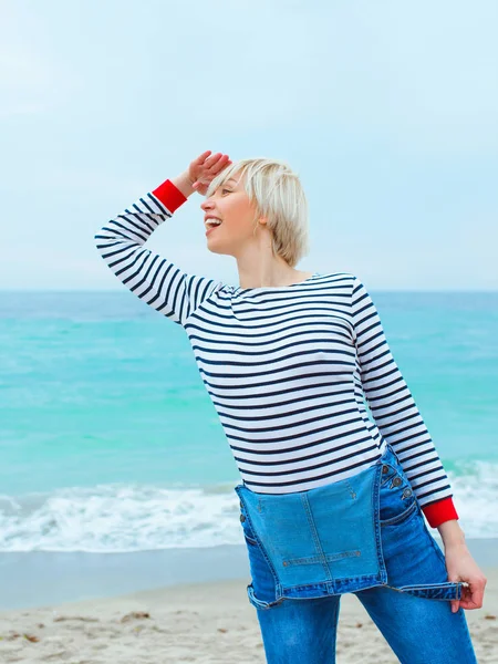 beautiful cute blonde woman sailor in striped blouse and jeans standing on the coast by the sea (ocean) and looking for