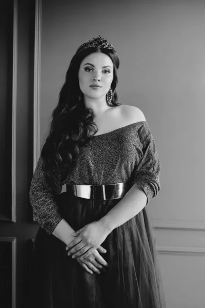 monochrome (black and white) portrait of beautiful young pretty cute woman princess (queen) in long purple queen\'s dress and crown, with long hair and make up indoor in minimalistic interior