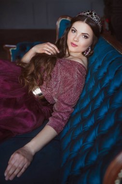 uzun saçlı uzun mor kraliçe elbise ve taç güzel genç güzel sevimli kadın prenses (kraliçe) portresi ve minimalist iç kapalı mavi kadife kanepede oturan makyaj