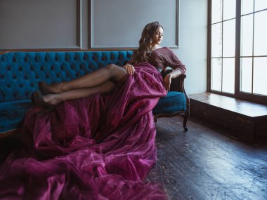 uzun saçlı uzun mor kraliçe elbise ve taç güzel genç güzel sevimli kadın prenses (kraliçe) portresi ve minimalist iç kapalı mavi kadife kanepede oturan makyaj