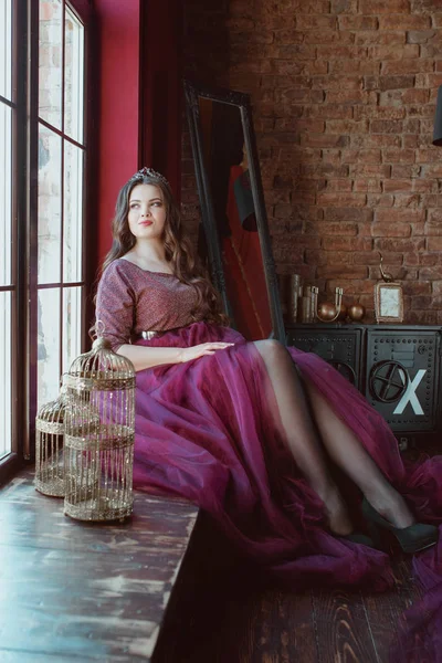 肖像美丽的年轻漂亮可爱的女人公主 在长紫色女王的礼服和皇冠 长头发和化妆室内在简约的阁楼内部坐在窗口 — 图库照片