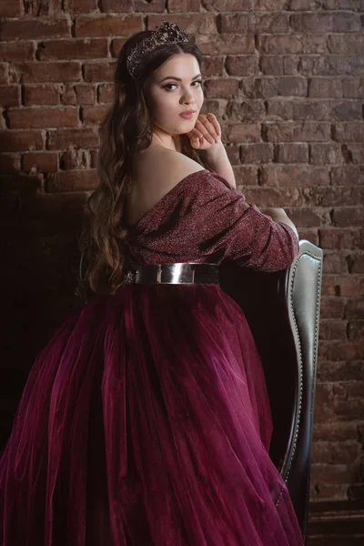 uzun saçlı uzun mor kraliçe elbise ve taç güzel genç güzel sevimli kadın prenses (kraliçe) portresi ve deri sandalye yakınında minimalist çatı iç kapalı makyaj
