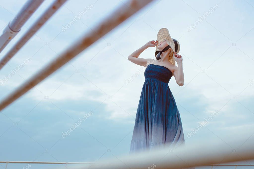 Caucasian beautiful stylish blonde woman in blue dress and in straw hat standing near the sea 