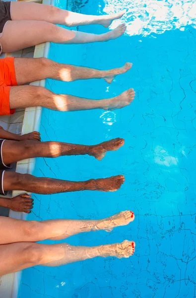 legs of four friends (common people) different race sitting by the swimming pool in sunny day. Party. Summer. Vacation and sport concept.
