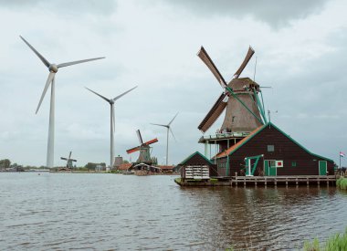 Bulutlu bir günde eski rüzgar değirmenleri ve modern rüzgar değirmenleri (Rüzgar jeneratörleri) ile Hollanda eski köy Zaanse Schans Kolaj. Eski ve yeni dönem, yüzyıl arasındaki kontrast. 2019 yılının baharı