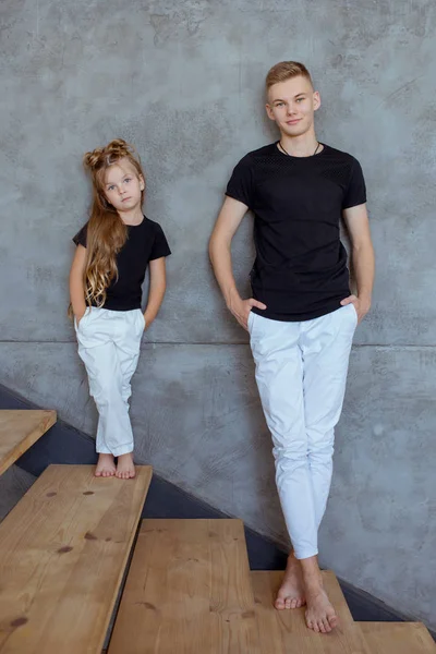 穿着深色T恤衫和白色裤子的时髦的高加索兄弟姐妹 站在阁楼内部灰色的墙壁旁边 关系和儿童概念 — 图库照片