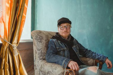 portrait of modern redhead handsome stylish man in black hat and Jean jacket and sitting in the armchair. Fashion and style concept clipart