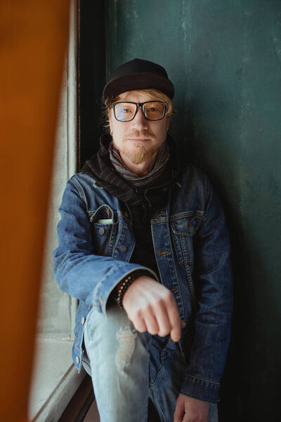 portrait of modern redhead handsome stylish man in black hat and Jean jacket and glasses alone by the window. Fashion concept
