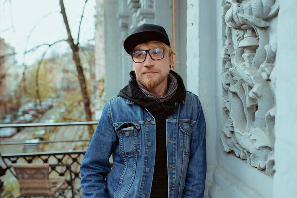 portrait of modern redhead handsome stylish man in black hat and Jean jacket and glasses standind on the balcony in old part of the city. Urban and fashion concept