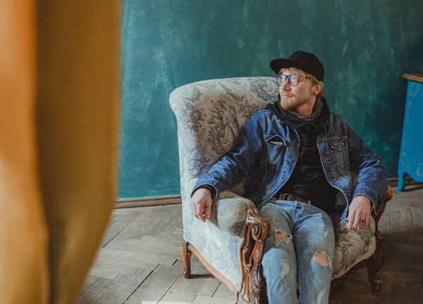 portrait of modern redhead handsome stylish man in black hat and Jean jacket and sitting in the armchair. Fashion and style concept