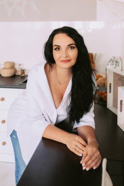 Mutfakta beyaz gömlek ve kot pantolon giyen esmer, şık bir kadın. Aşçılık ve moda konsepti