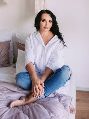 Beyaz gömlekli, kot pantolonlu, yatağında üzgün oturan güzel siyah saçlı kadının portresi. Duygular, kriz, ev, iç konsept