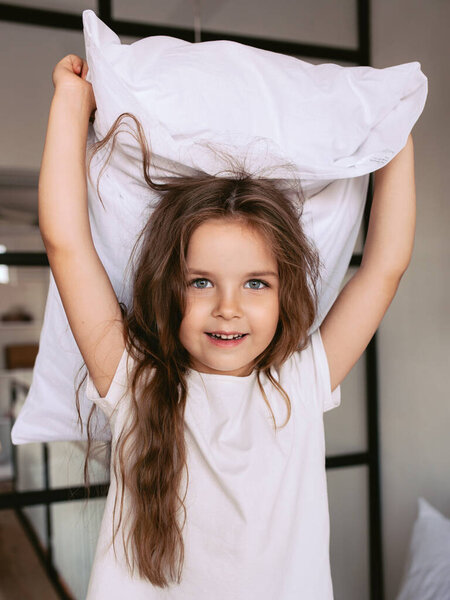 Adorable cheerful little girl in white t-shirt fighting pillow at home indoor. Fashion, fun, style, childhood, emotions, growing up concept