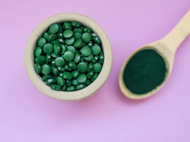 Masa kaşıklarıyla yeşil Hawaii spirulinası hapları ve açık leylak arka planda pudra. Süper yemek, sağlıklı yaşam tarzı, sağlıklı destek konsepti.