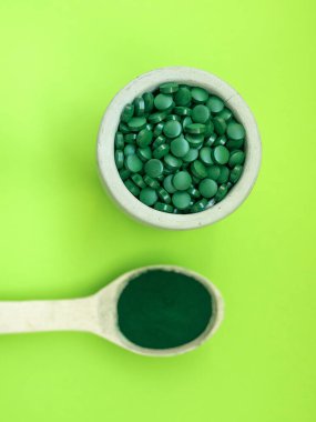 Masa kaşıklarında yeşil spirulina hapları ve açık yeşil arka planda toz. Süper yemek, sağlıklı yaşam tarzı, sağlıklı destek konsepti.