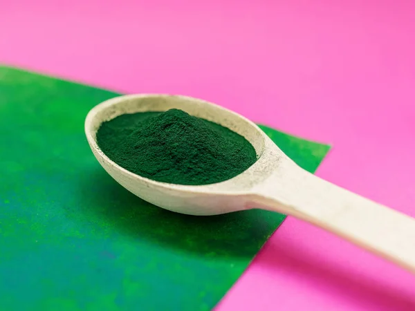 Green hawaiian spirulina in powder in wooden spoon on pink and green background. Super food, healthy lifestyle, healthy supplements concept