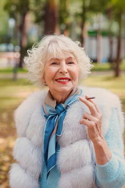 smiling senior elegant stylish fashionable woman with grey hair in fur coat outdoor smoking cigarette. Unhealthy lifestyle, age, positive vibes, oldness, addiction, bad habit concept.