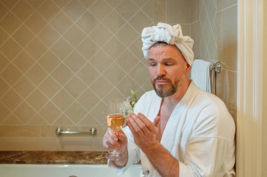 Beyaz bornozlu ve kafasında havluyla banyoda köpükle oturmuş gül şarabı içen yakışıklı olgun bir adam. Spa, rahatla, yaşam tarzı, hayatın tadını çıkar.