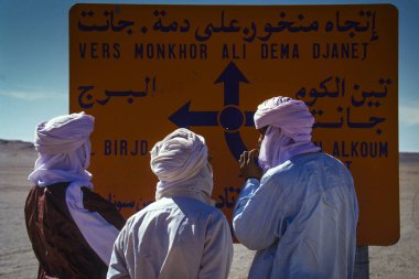TASSILI NAJJER, ALGERIA - JANUARY 10, 2002: Unknown men check the road sign with their off-road vehicles in the sand dunes of the Algerian Sahara desert, Africa, Tassili NAjjer National Park clipart