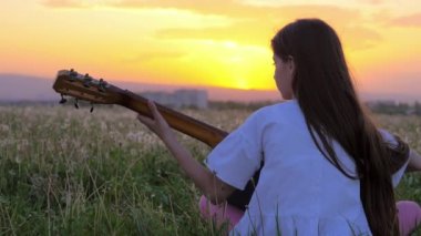 sırtını bir karahindiba alandaki gitar çalmak kamera ile oturan güzel günbatımı küçük çocuk bir kız. Açık havada şehir binaları hippi tarzı uzak