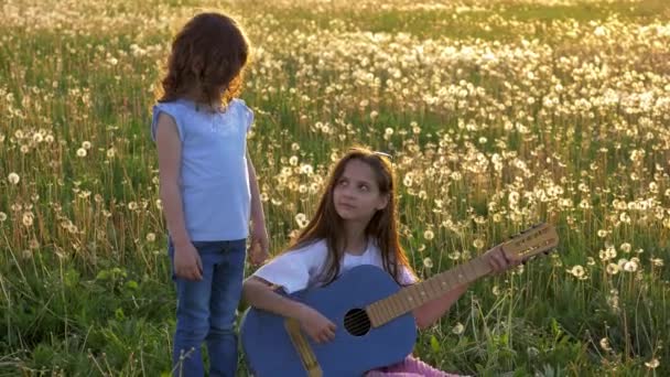 两个滑稽的姐妹在蒲公英地里唱歌和演奏声学牛仔裤吉他 在户外娱乐休闲的小女孩 — 图库视频影像